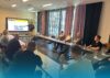 mehrere Personen sitzen an einem U-förmigen Tisch in einem Meetingraum und schauen auf eine digitale Tafel und hören einem Mann zu