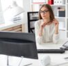 Frau sitzt an einem hellen Büroarbeitsplatz mit Computer und Tastatur und schaut auf einen Bildschirm